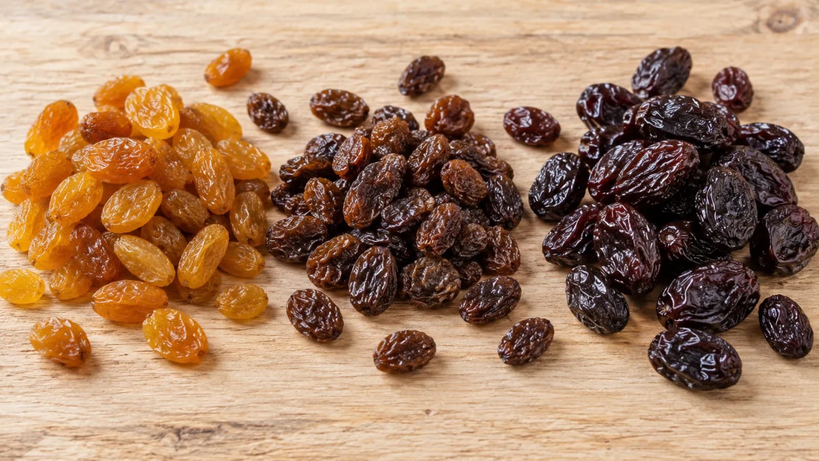 Pasas de uva: Flame, Sultanina o Thompson; Argentina, Chile o Turquía, y ocratoxina A Tres montones de pasas de uva sin semilla sobre una tabla de madera clara: Sultanina dorada, Thompson Seedless marrón oscura y Flame Seedless rojiza casi negra, de mayor calibre